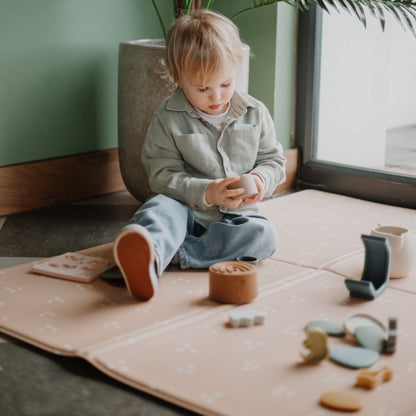 Child playing with toy silicone toys on a mat in a room with plants and a window.
