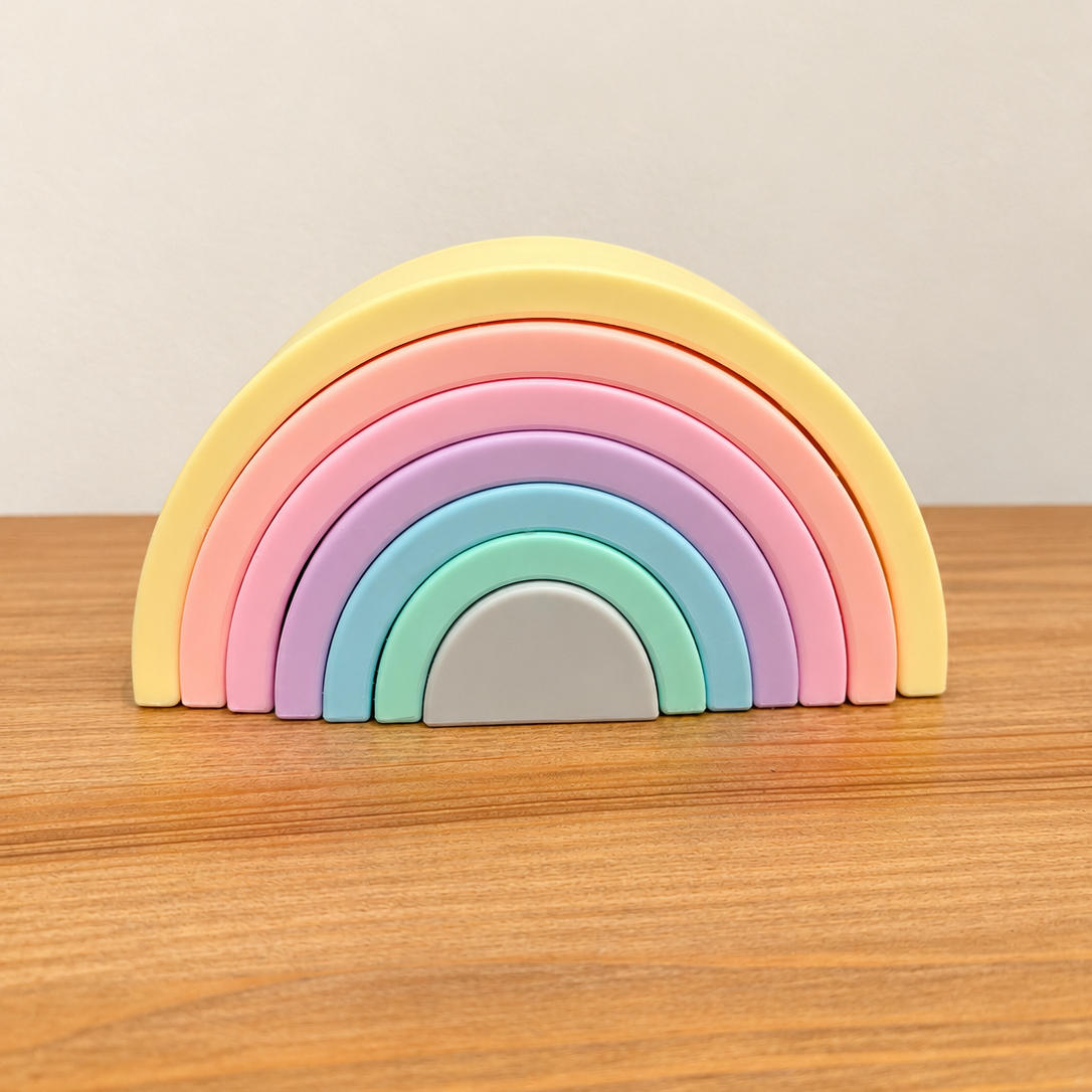 A pastel rainbow silicone toy with five semi-circular arches in yellow, orange, pink, purple, and light blue, with a small white arch at the base, resting on a wooden tabletop against a light beige wall.