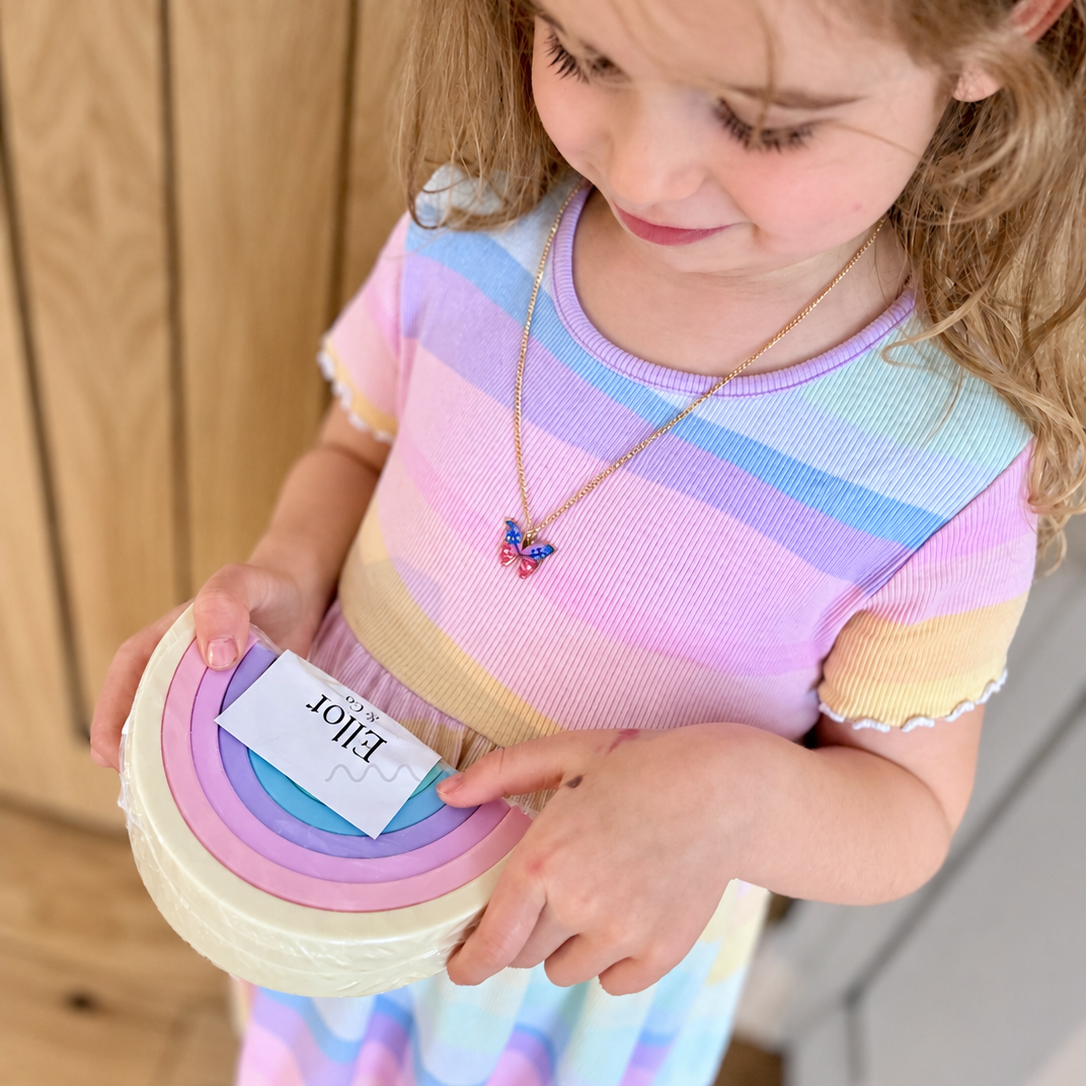 A young girl with blonde hair wearing a pastel rainbow striped shirt holds a stack of pastel rainbow silicone stacking toy, pointing to a block labeled ellor and co